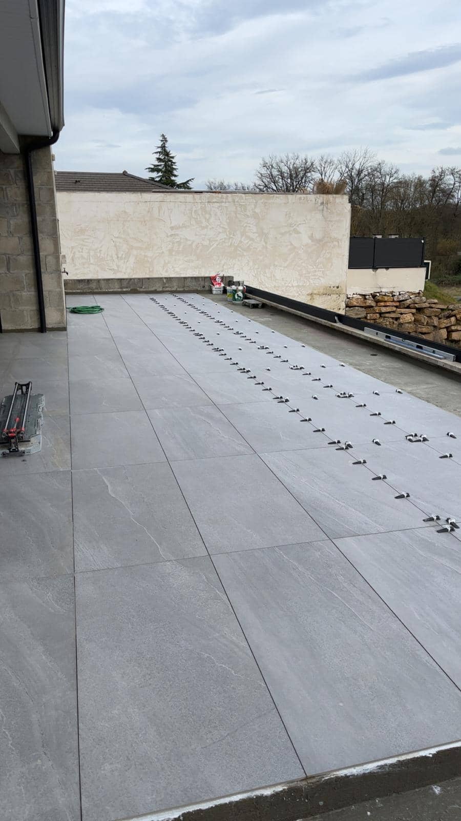 Pose de grandes dalles de grès cérame gris effet pierre sur une terrasse, clips et cales de nivellement sur les derniers rangs, coupe-carreau manuel au premier plan, réalisation à Rumilly