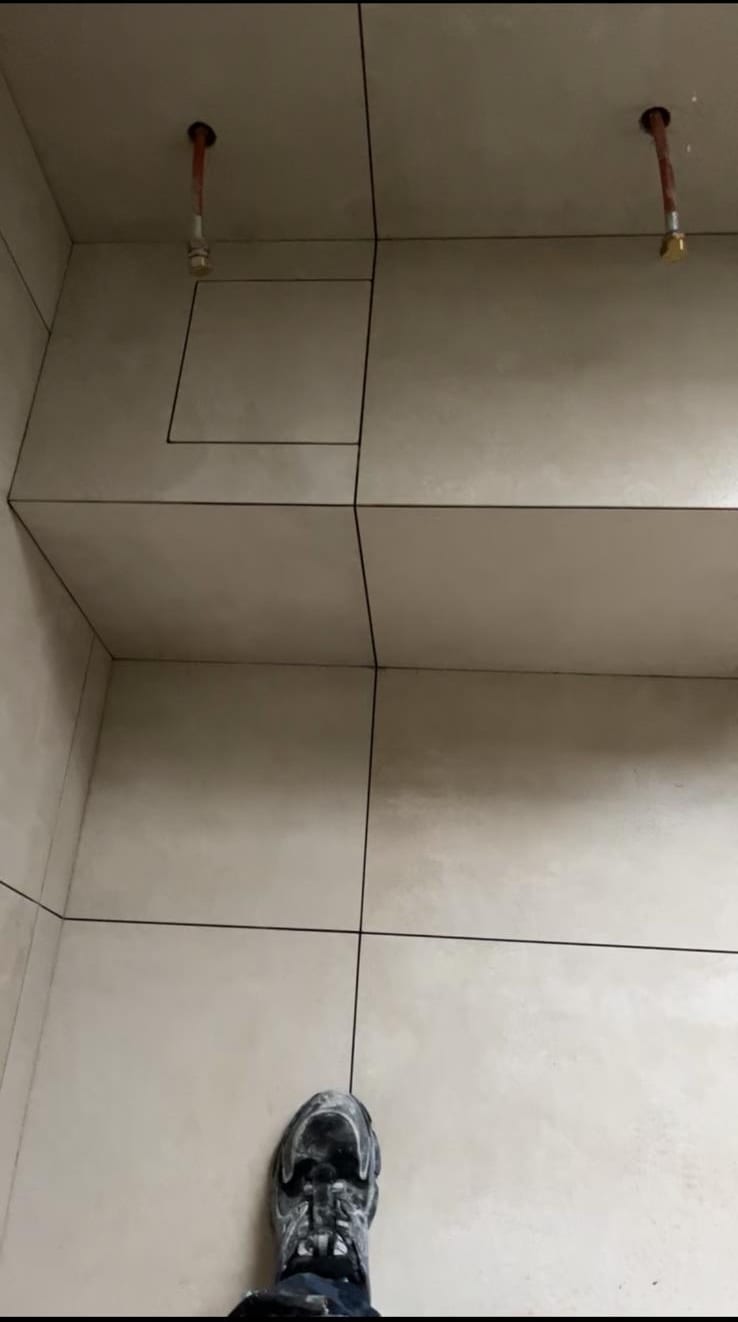 Vue plongeante sur un sol et un podium en grands carreaux beige à joints fins foncés alignés, trappe de visite intégrée, chaussure d’ouvrier au premier plan, réalisation à Chambéry