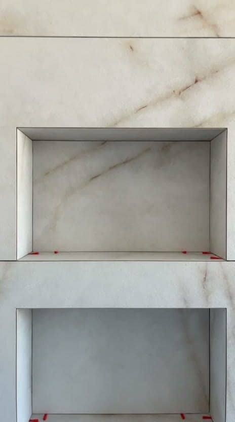 Deux niches superposées habillées de grès effet marbre blanc veiné beige, arêtes nettes sans profilé, cales rouges encore posées sur les tablettes, réalisation à Thonon-les-Bains