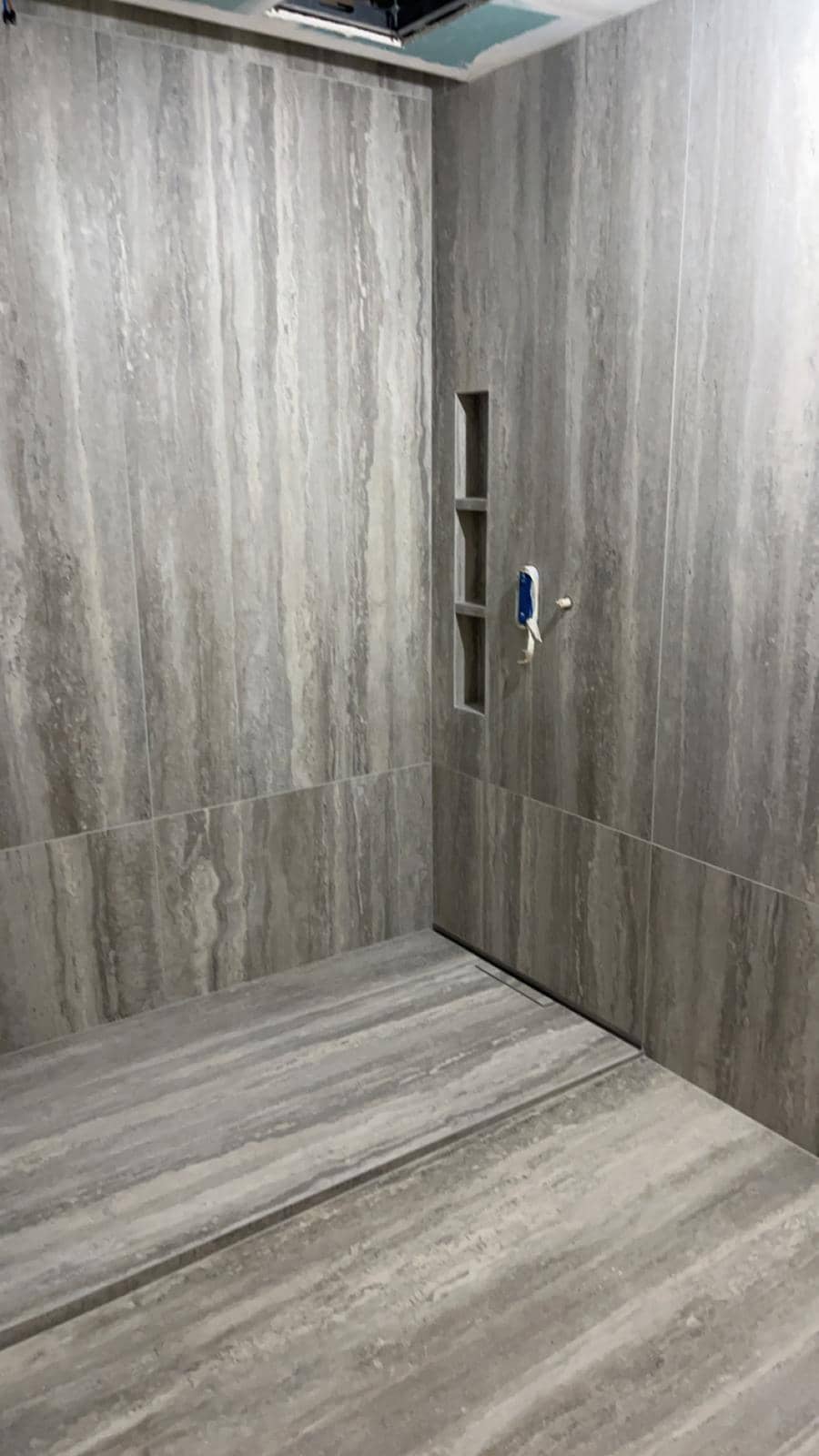 Douche de plain-pied en grès cérame très grand format effet travertin gris, veinage continu d’une plaque à l’autre, caniveau linéaire et niche verticale à trois compartiments, réalisation à Annecy