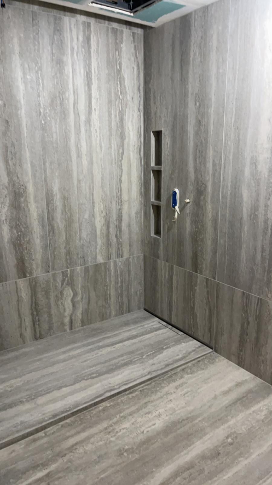Douche de plain-pied en grès cérame très grand format effet travertin gris, veinage continu d’une plaque à l’autre, caniveau linéaire et niche verticale à trois compartiments, réalisation à Annecy